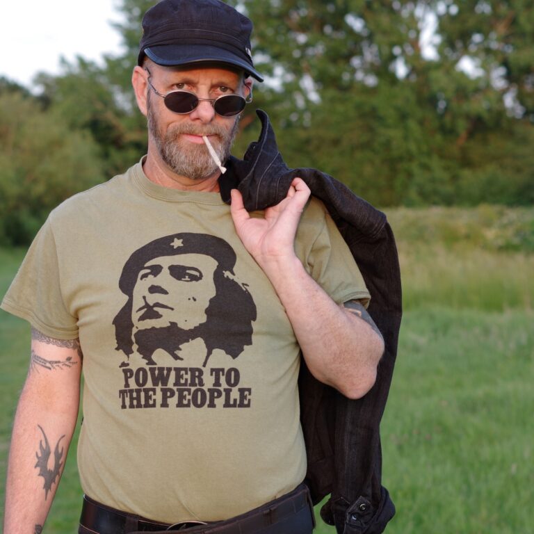 Stuart Welbourn standing outdoors at golden hour, wearing a Citizen Smith t-shirt and black denim jacket, holding the jacket over his shoulder with a defiant expression.